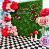 Enchanted Wonderland Set featuring a lush green foliage wall, colourful balloon garland in red, pink, lavender, and blue, oversized red, blush pink, and purple roses, decorative gold frames, red roses, whimsical mushrooms, and a black-and-white checkered floor. Alice in Wonderland birthday party backdrop by Magic Flowers Event Rentals in Toronto and GTA.