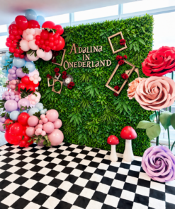 Enchanted Wonderland Set featuring a lush green foliage wall, colourful balloon garland in red, pink, lavender, and blue, oversized red, blush pink, and purple roses, decorative gold frames, red roses, whimsical mushrooms, and a black-and-white checkered floor. Alice in Wonderland birthday party backdrop by Magic Flowers Event Rentals in Toronto and GTA.