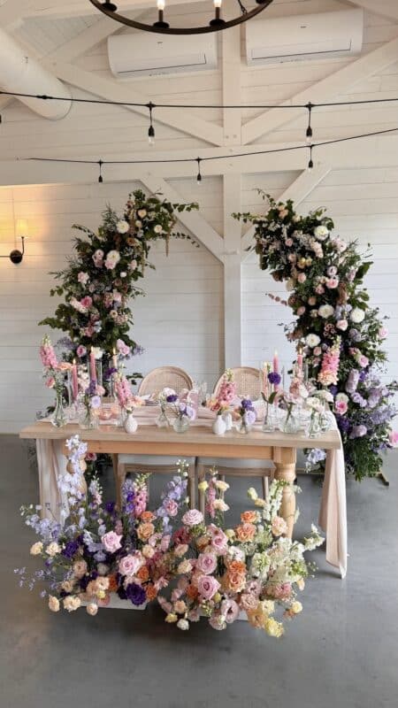 Elegant wedding table setup with pastel flower arrangements, floral arch backdrop, and romantic decor in a white barn-style venue