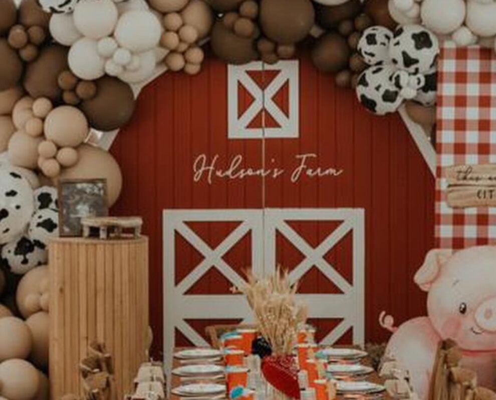 Custom farm-themed kids birthday party setup in Toronto featuring a red barn house backdrop, 'Hudson's Farm' signage, table setup, wooden plinth, shelved treat backdrop and a cow-print balloon arch by Magic Flowers Event Rentals.