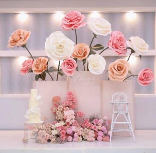 A luxury baptism decor setup featuring giant handcrafted roses in shades of blush pink, cream, and peach, arranged in large minimalist vases. The display includes a matching floor-level floral arrangement, a white tiered cake, and a vintage-style high chair.