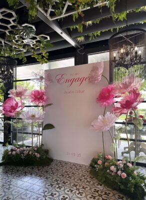 Large white square engagement backdrop with "We are Engaged" script and oversized pink and white handcrafted flowers for an event rental in Richmond Hill.
