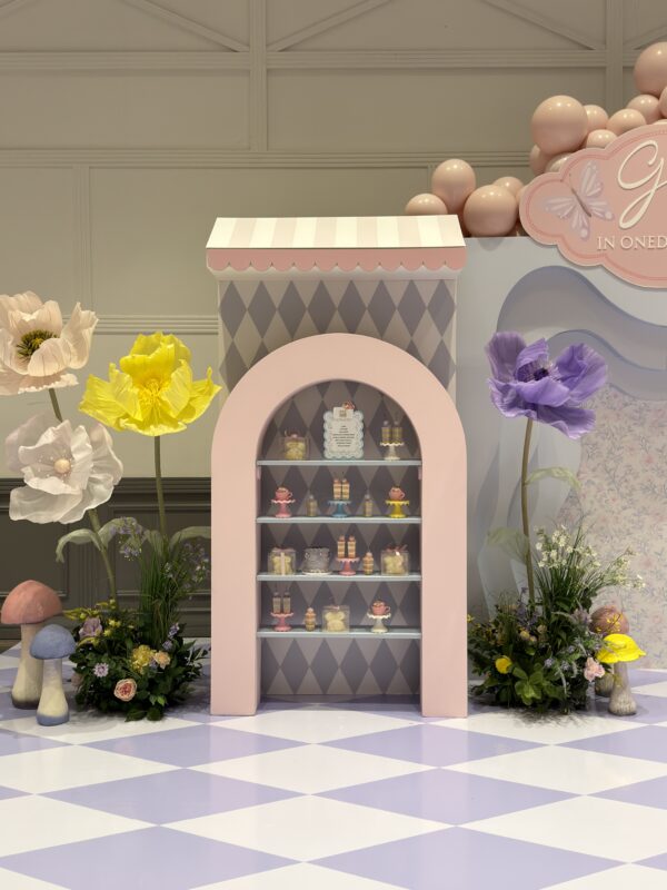 Whimsical Alice in Onederland 1st birthday decor setup at Embassy Grand Brampton, featuring a custom storybook backdrop, a giant standing flower garden, and a vintage-style blue treat cart surrounded by pastel balloon clusters on a lavender checkered floor.