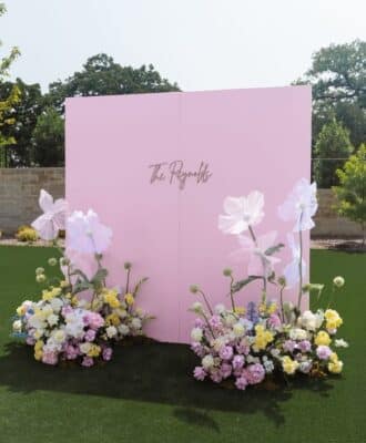Large pink square backdrop with custom gold signage and white floral accents for an outdoor event rental in Newmarket.