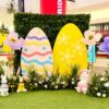 A wide view of an Easter event decor setup at a mall, featuring a greenery wall backdrop, two giant decorative eggs, large artificial spring flowers, and three bunny statues sitting on a green turf floor.