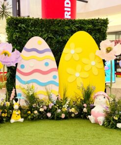 A wide view of an Easter event decor setup at a mall, featuring a greenery wall backdrop, two giant decorative eggs, large artificial spring flowers, and three bunny statues sitting on a green turf floor.