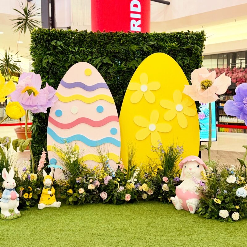 A wide view of an Easter event decor setup at a mall, featuring a greenery wall backdrop, two giant decorative eggs, large artificial spring flowers, and three bunny statues sitting on a green turf floor.