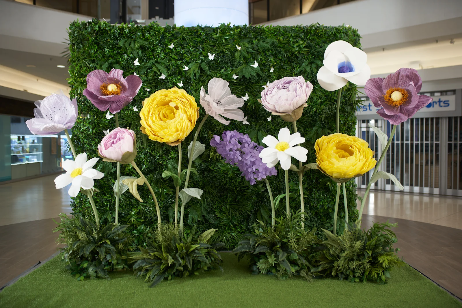 Large boxwood greenery wall backdrop decorated with small white butterflies, featuring a vibrant display of oversized freestanding flowers in yellow, purple, pink, and white. Set up on artificial grass as an Easter holiday photo stage inside Bridlewood Mall in Scarborough.