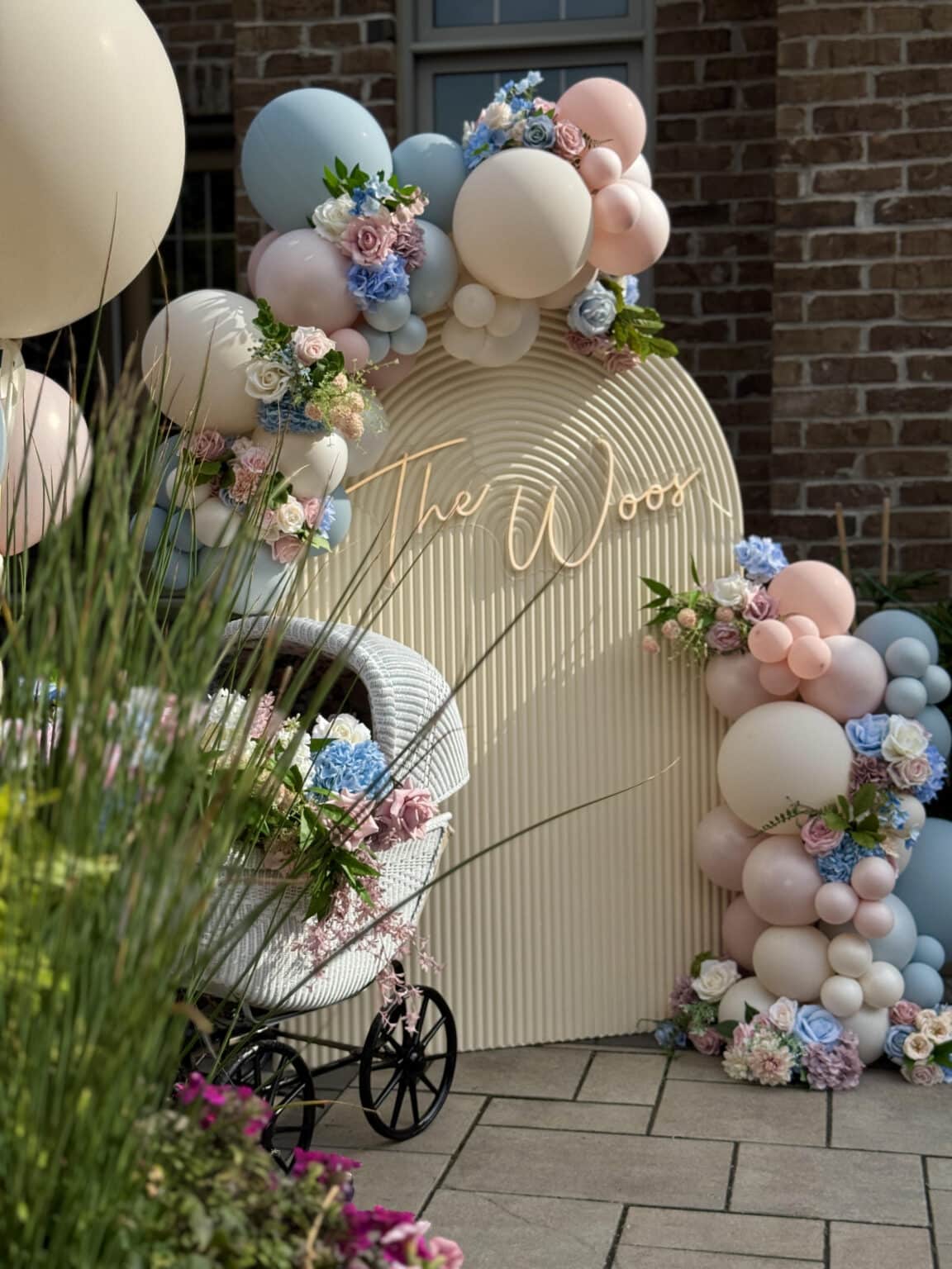 Outdoor baby shower setup in Markham featuring a beige ribbed arch backdrop with custom signage, decorated with a pastel balloon and floral garland, next to a vintage white wicker baby carriage filled with flowers.