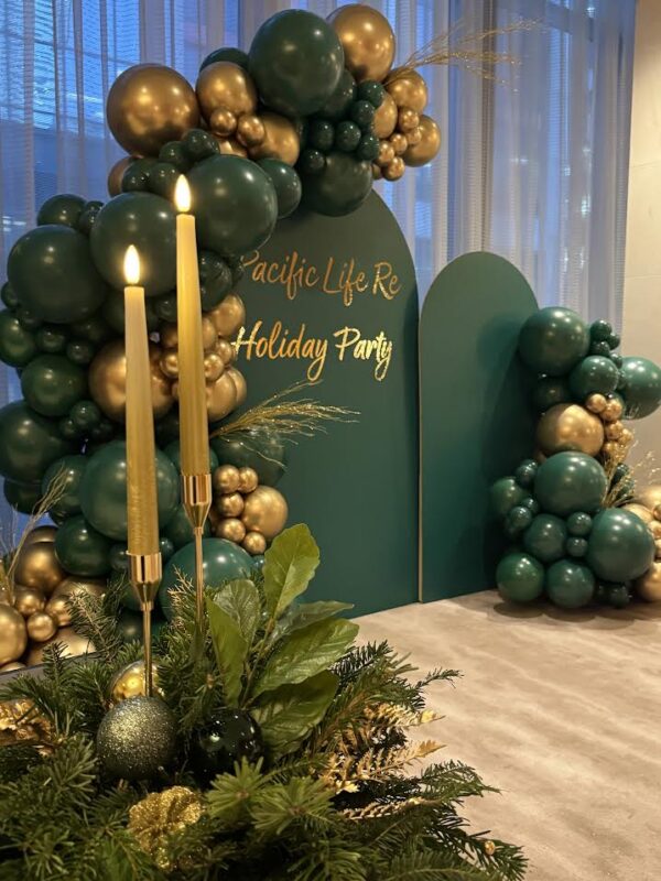 Emerald green and metallic gold balloon garland draped over two custom green arched backdrops with gold text reading 'Pacific Life Re Holiday Party'. The foreground features a festive evergreen centerpiece with ornaments and two tall gold taper candles.