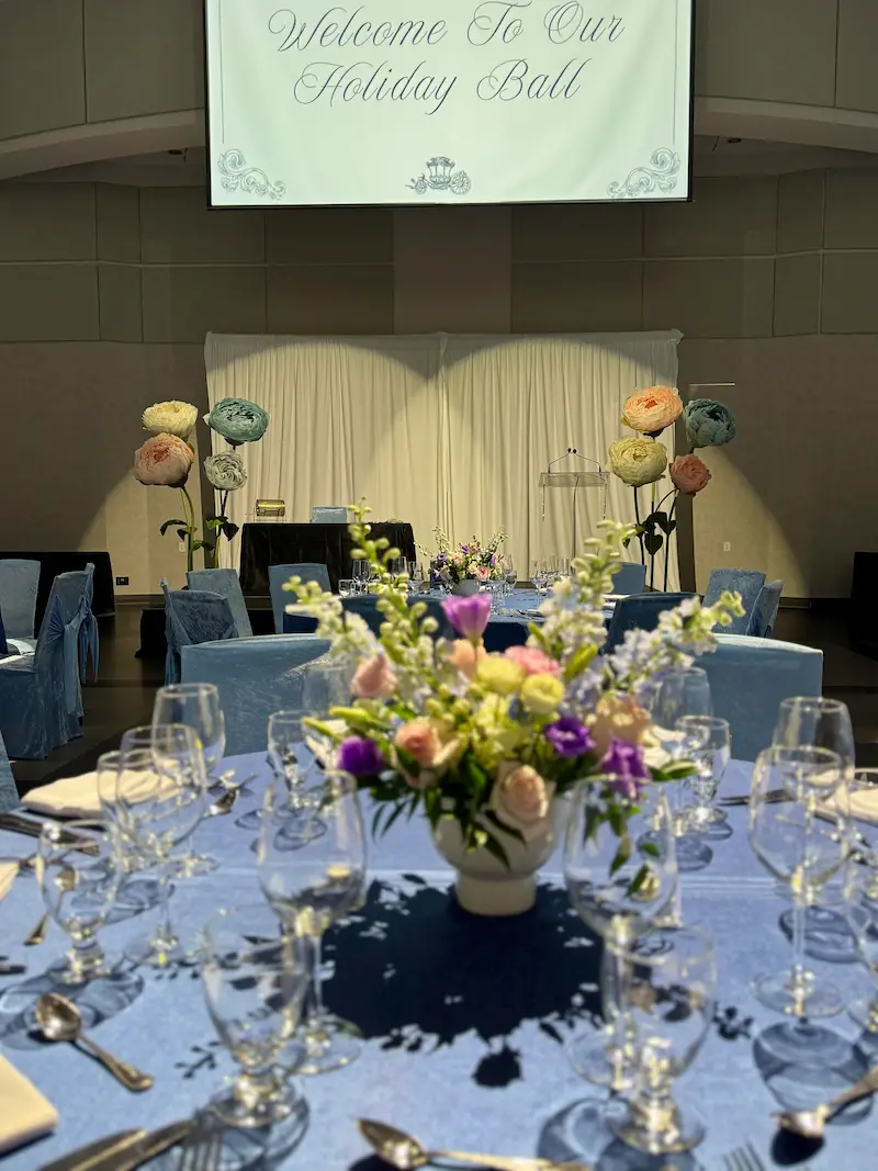 The set up for a corporate holiday ball at Paramount Event Space in Woodbridge, Vaughan, featuring blue table linens, floral centerpieces, and giant standing flower props on the stage.