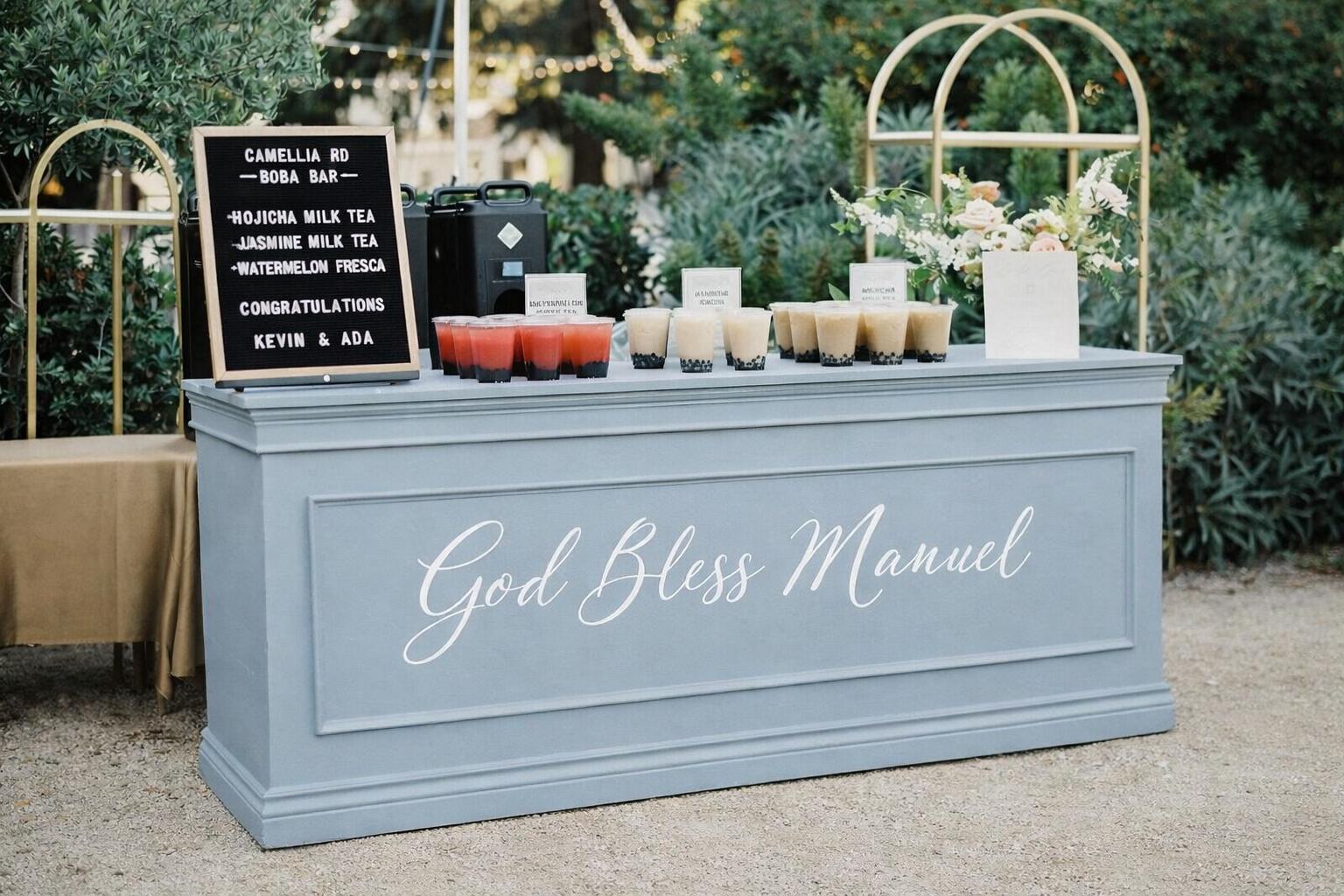A customized light blue paneled dessert table setup for an outdoor event. The front of the table features a large white script decal that says "God Bless Manuel". On the table are assorted beverages, a black letterboard sign, and a small floral arrangement with white and pink flowers.