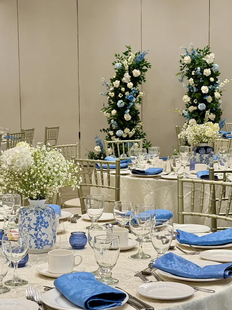 A banquet hall event decor setup at the Mississauga Convention Center featuring baby's breath floral table centerpieces in blue and white vases, silver chairs, and two tall blue and white flower cascades in the background.