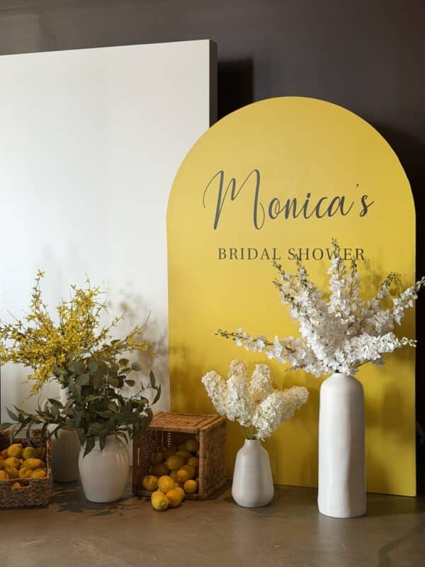 Custom yellow arched bridal shower backdrop with white Mediterranean-style vases, tall floral arrangements, and woven baskets filled with fresh lemons.