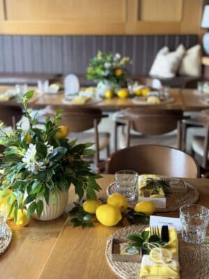Wooden table set for a lemon-themed bridal shower featuring woven hyacinth placemats, yellow napkins, and a ribbed white ceramic vase centerpiece with greenery and lemons.