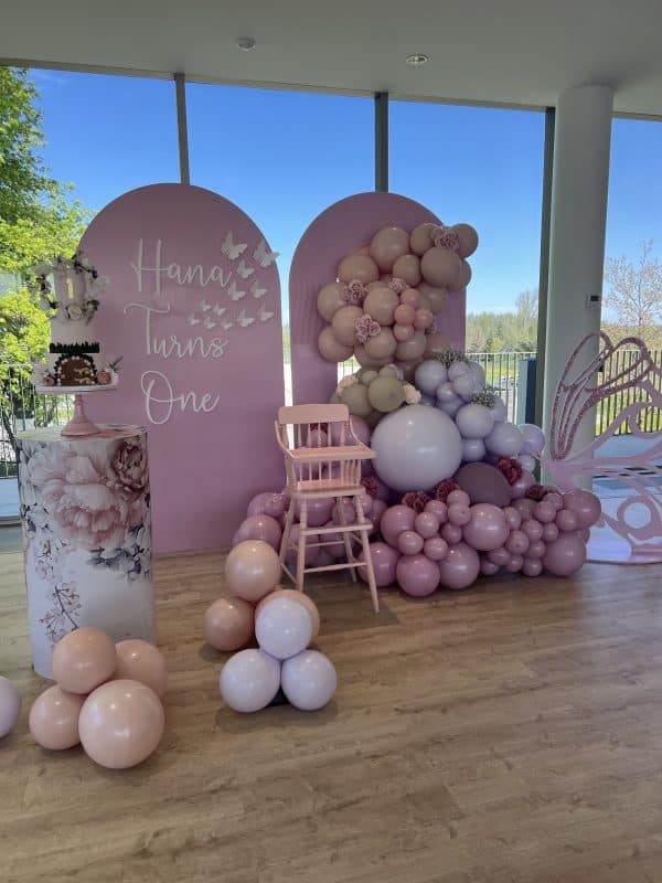 Pink butterfly-themed first birthday party setup with arched backdrops, pastel balloon garlands, and a floral cake pedestal.