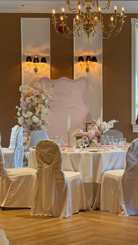 Pastel pink wavy backdrop with Baby in Bloom sign, framed by lush floral cascades and elegant table centerpieces at The School Fine Dining in Markham.