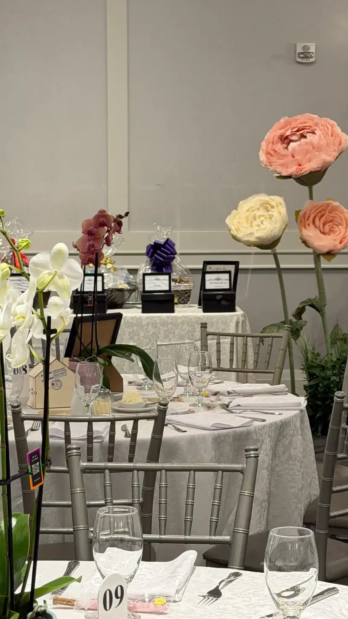 Three large, freestanding oversized flowers in shades of pink and cream stand behind a formally set banquet table featuring silver Chiavari chairs and potted orchid centerpieces at Parkview Manor.