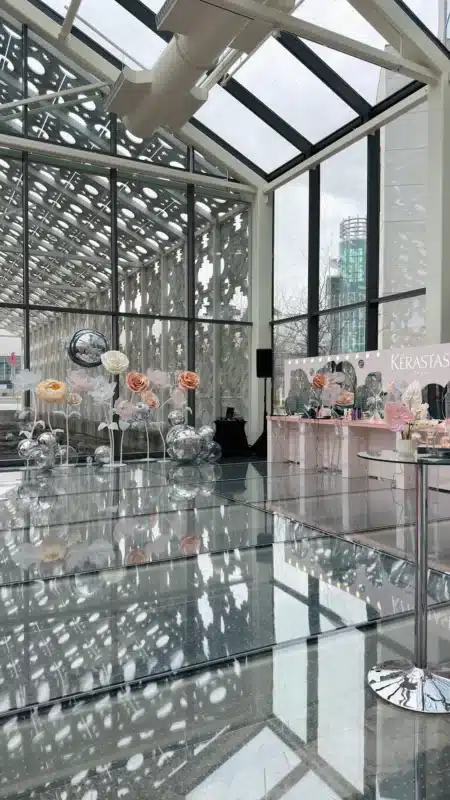 A bright, glass-enclosed event space at Hotel X in Toronto featuring a pink Kérastase styling vanity, decorated with giant pastel silk flowers and decorative silver disco balls.