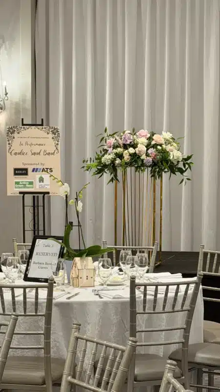 A tall golden plinth topped with an oversized arrangement of pink, white, and lavender flowers stands beside a formally set dining table at the St. Clare Inn Spring Fundraiser in Parkview Manor.
