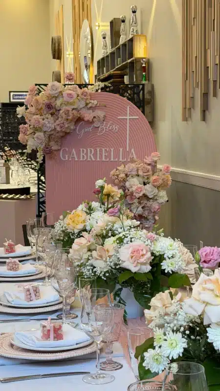 A beautifully decorated banquet table featuring lush pink and peach floral centerpieces and a custom pink arched backdrop reading 'God Bless Gabriella' for a spring baptism event at Trattoria Tovico in Vaughan.