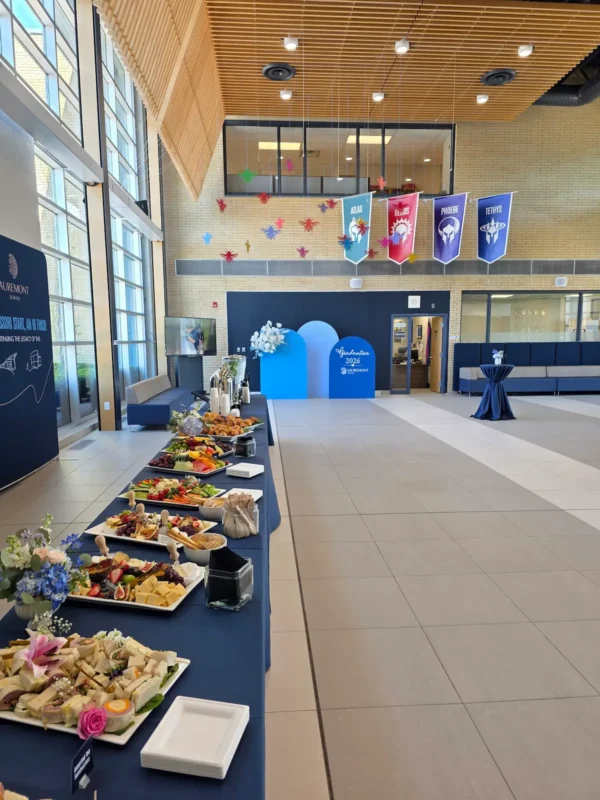 A bright event space featuring a dark and light blue arched backdrop setup, decorated with white and blue floral arrangements and a custom white decal reading "Graduation 2026 Lauremont School".
