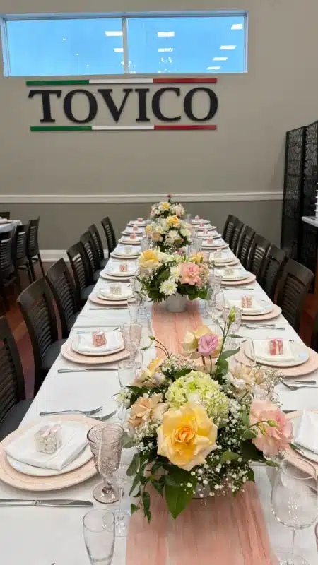 A long dining table set for a baptism at Trattoria Tovico in Vaughan, featuring pastel floral centerpieces by Magic Flowers Event Rentals, pink sheer table runners, textured charger plates, and small party favors.