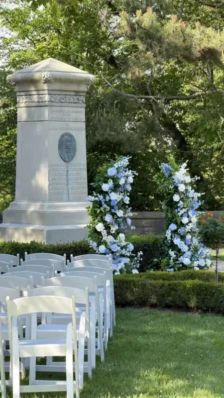 Rows of white folding chairs arranged on a green lawn facing two semi-circular floral cascades made of blue and white roses, positioned next to a stone monument for an outdoor wedding ceremony.