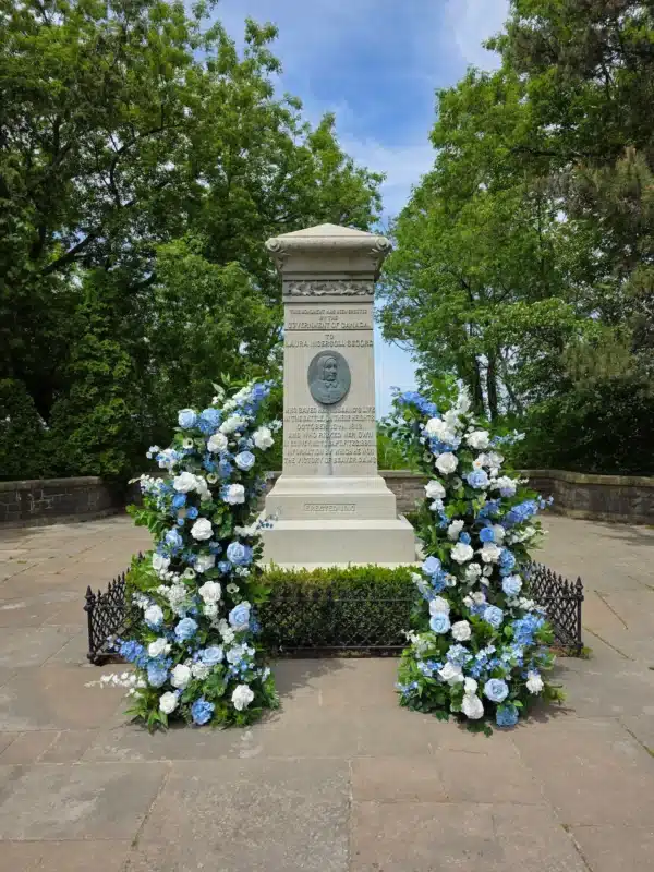 Rows of white folding chairs arranged on a green lawn facing two semi-circular floral cascades made of blue and white roses, positioned next to a stone monument for an outdoor wedding ceremony.
