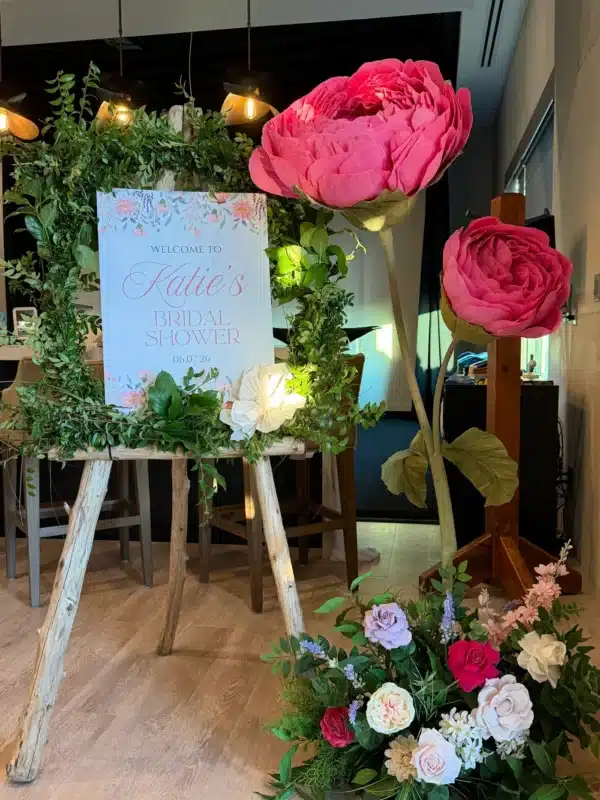 A rustic wooden easel holding a welcome sign for Katie's Bridal Shower, draped in a lush greenery wreath. Next to it stands a vibrant pink, large standing flower on a wooden base, surrounded by a floor arrangement of mixed roses and greenery