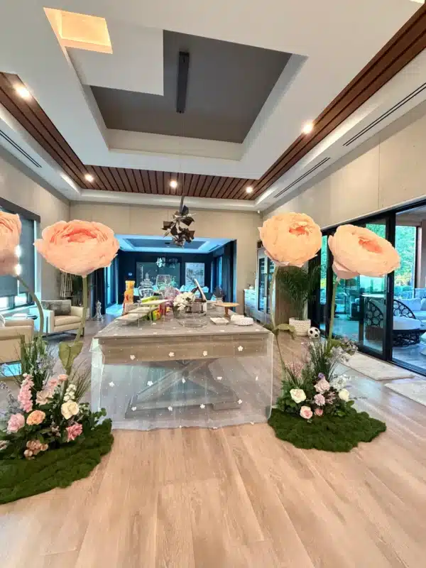 A modern dining space set up for a bridal shower, featuring a rustic wooden table draped with a sheer, white dotted overlay. The table is flanked by two giant, freestanding light pink flowers with green stems, resting on decorative moss patches mixed with smaller, colorful floral arrangements.