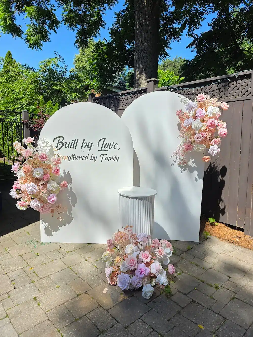 A photo of a beautiful, outdoor event decor setup on a stone patio in Oakville for a marriage anniversary. The setup features two large, interlocking white arched backdrop panels. On the left panel, the message "Built by Love, Strengthened by Family" is written in elegant gold lettering. A fluted white column pedestal stands between them. Three abundant floral arrangements of blush pink, white, and peach roses and greenery are placed on the arches and the ground. The background features a dark wood fence and lush green trees under a clear blue sky.