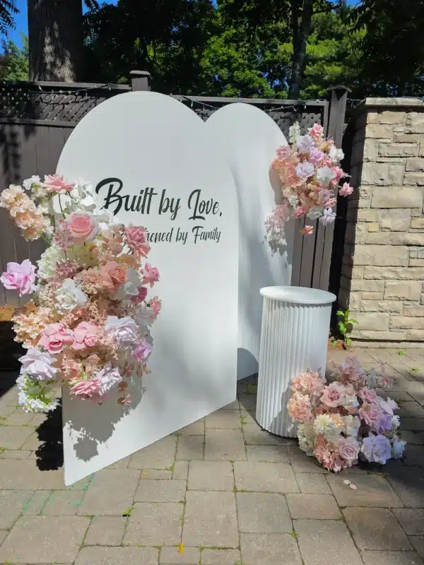 A close-up view of a white arched event backdrop on an outdoor stone patio featuring the custom text "Built by Love, Strengthened by Family." The arch is decorated with cascading blush pink, peach, and white floral arrangements. A white fluted pedestal stands on the right alongside a matching ground-level floral arrangement.