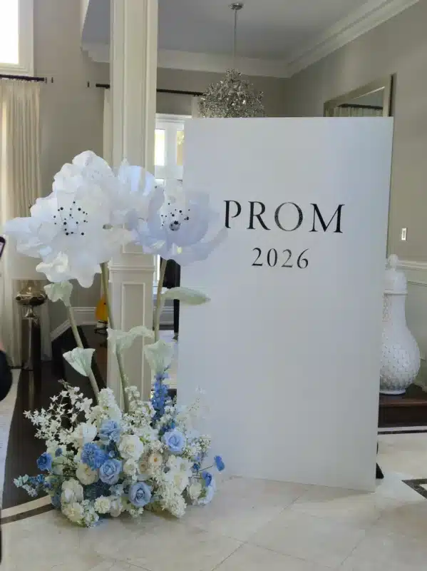 A white rectangular backdrop reading PROM 2026, decorated with oversized white artificial flowers on tall stems and a base floral arrangement of light blue, cream, and white roses, set up in a Whitchurch-Stouffville home foyer.
