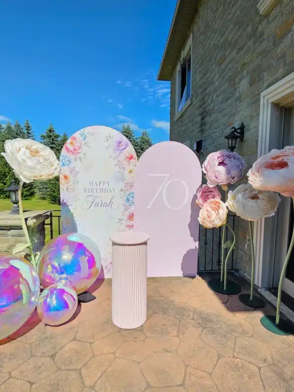 Outdoor 70th birthday party setup for "Farah" in Richmond Hill, Ontario, on a stone patio. Features custom-printed and pink arch backdrops, freestanding giant paper peonies, iridescent balloons, and a ripple fluted cake stand.