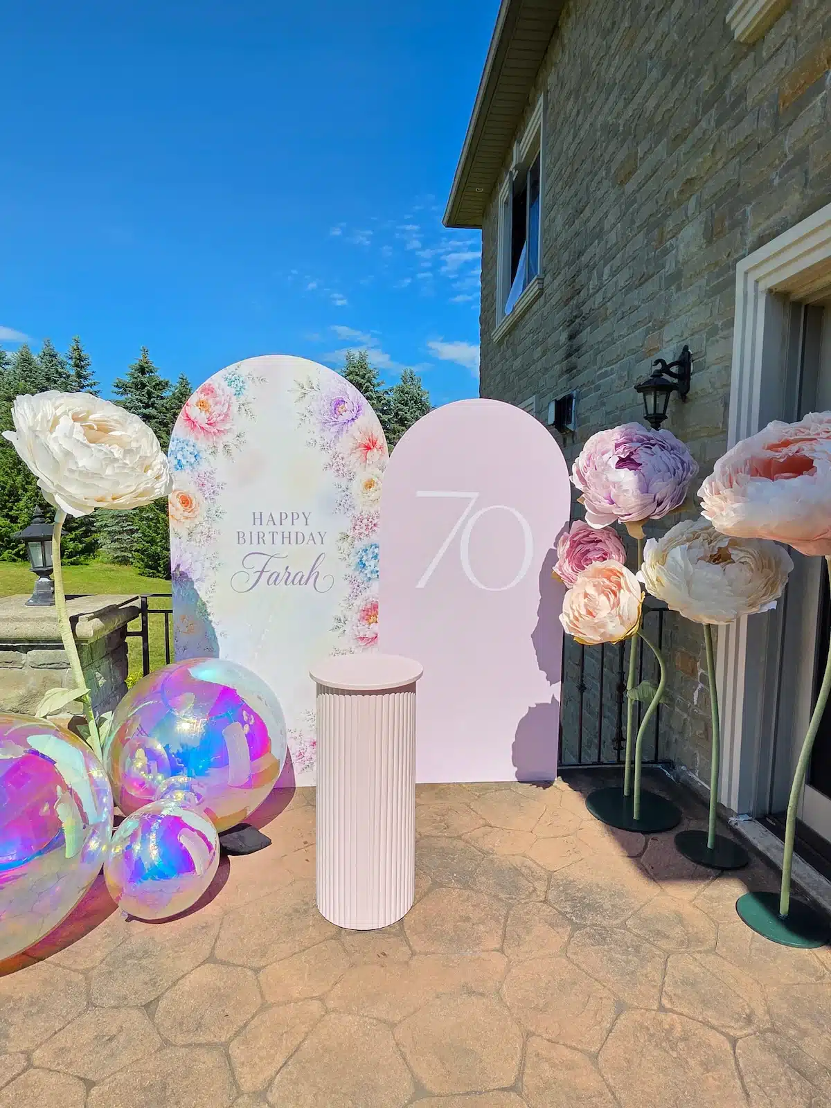 Outdoor 70th birthday party setup for "Farah" in Richmond Hill, Ontario, on a stone patio. Features custom-printed and pink arch backdrops, freestanding giant paper peonies, iridescent balloons, and a ripple fluted cake stand.