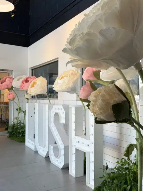 Giant freestanding pastel paper flowers and illuminated white marquee letters spelling LUSH, set against a modern tiled wall with greenery accents for a corporate brand activation.
