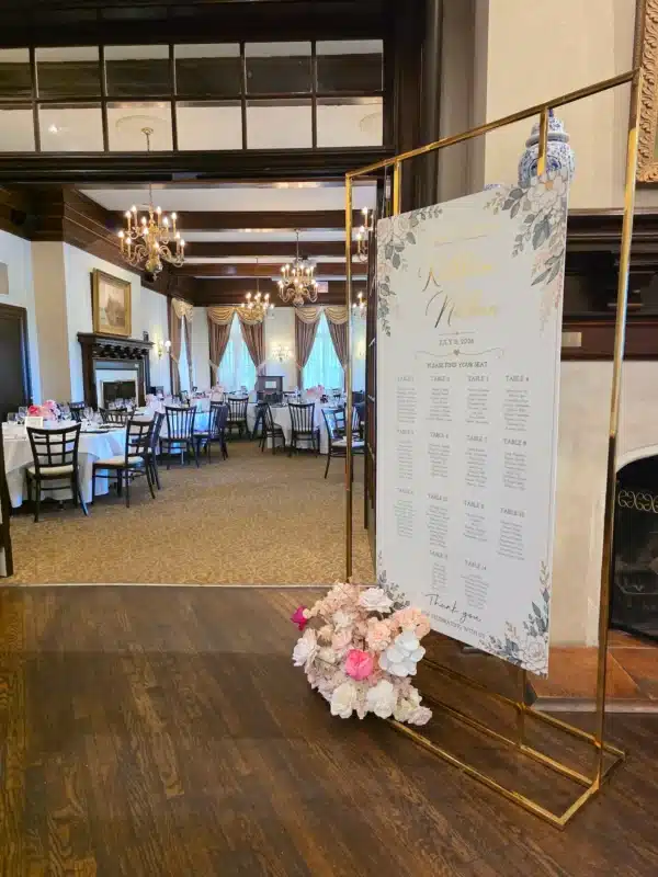 Custom floral wedding seating chart for Kathleen and Nathan suspended in a modern gold metal frame stand rental, accented with a base of pink and white roses, positioned at the reception entrance of Scarboro Golf Club.