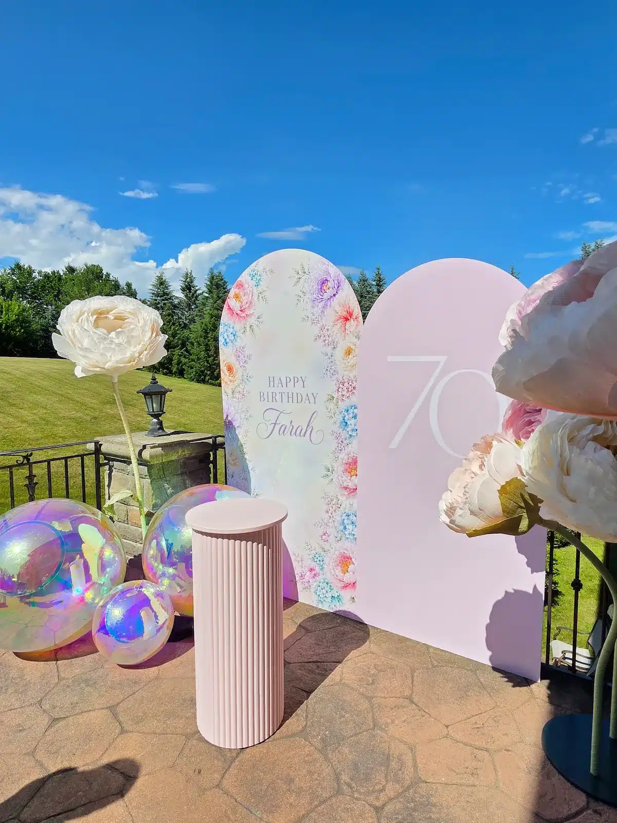 A full view of the personalized 70th birthday decor for Farah in Richmond Hill, featuring custom floral and milestone arch backdrops, giant freestanding paper flowers, iridescent balloons, and a fluted pink cake stand on a stone patio.