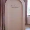 Alt Text Minimalist dusty-rose backdrop with an architectural arch shape, featuring black script text that reads "Hodman's BRIDAL BRUNCH," set against soft pink curtains and light wooden parquet flooring.