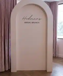 Alt Text Minimalist dusty-rose backdrop with an architectural arch shape, featuring black script text that reads "Hodman's BRIDAL BRUNCH," set against soft pink curtains and light wooden parquet flooring.