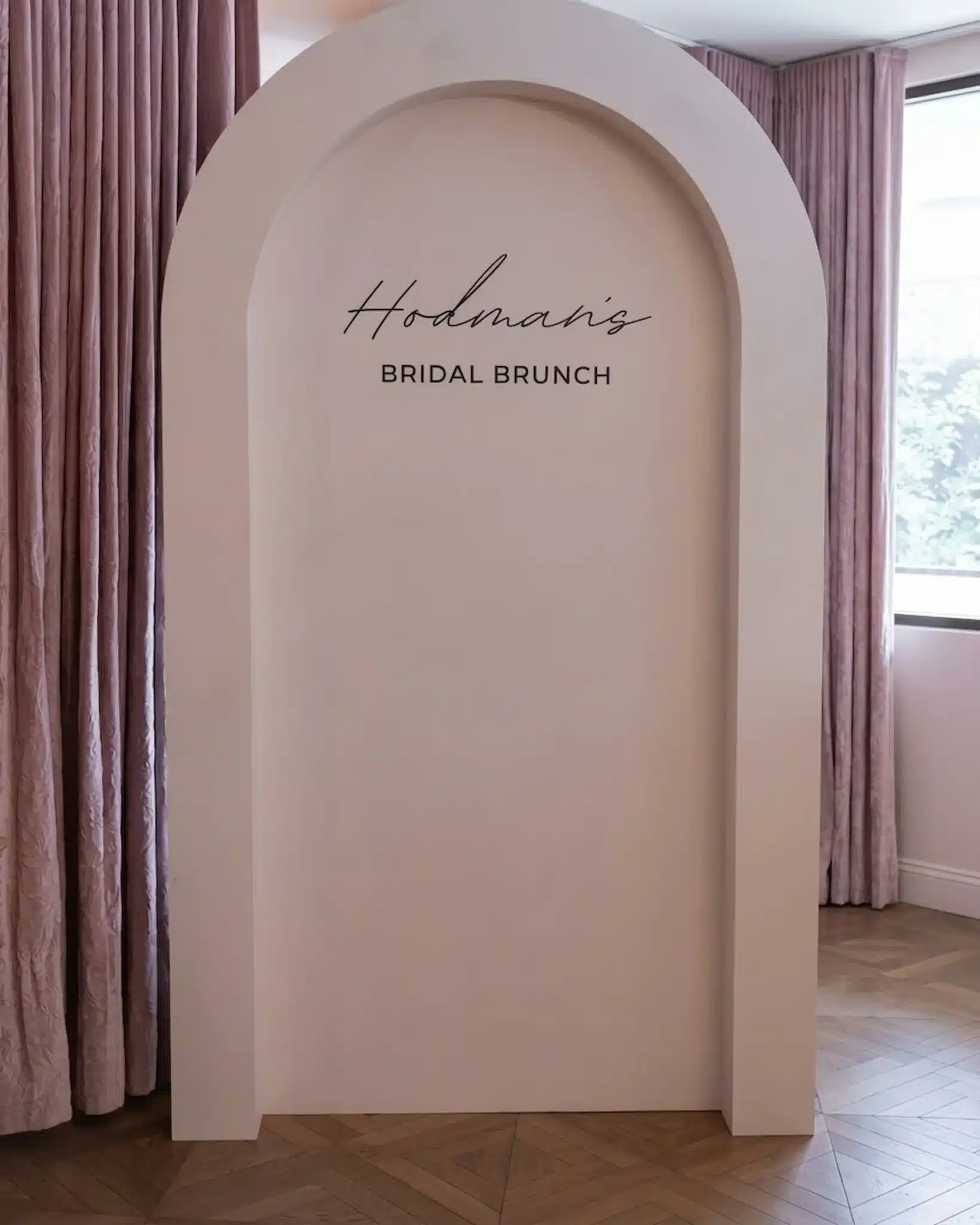 Alt Text Minimalist dusty-rose backdrop with an architectural arch shape, featuring black script text that reads "Hodman's BRIDAL BRUNCH," set against soft pink curtains and light wooden parquet flooring.