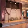 Elegant bridal brunch stage decor with layered blush backdrops, central arched panel, mauve draping, custom lettering, floral floor arrangement, acrylic podium, and cake pedestal.