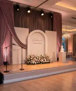 Elegant bridal brunch stage decor with layered blush backdrops, central arched panel, mauve draping, custom lettering, floral floor arrangement, acrylic podium, and cake pedestal.