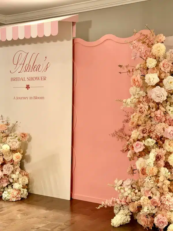 A detailed view of high-quality blush and cream silk flowers cascading down a wavy pink event wall, positioned next to a white panel featuring personalized bridal shower lettering.