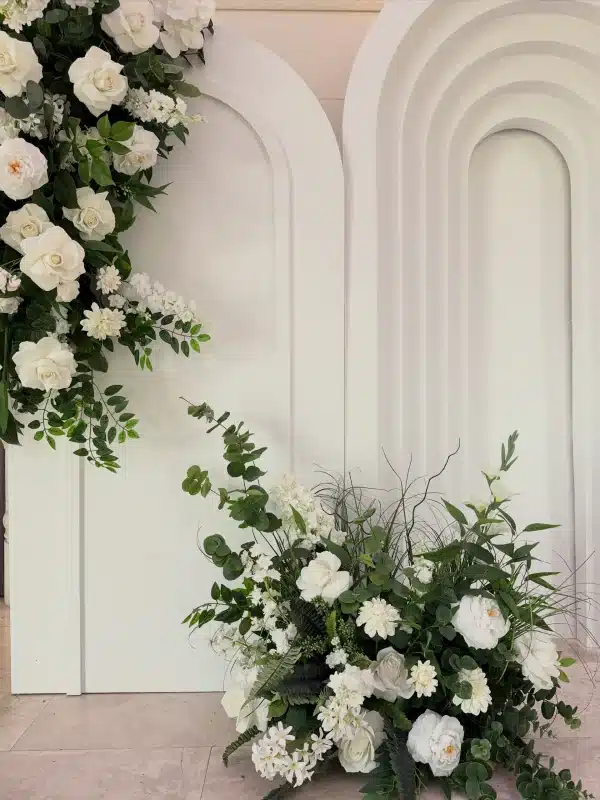 Close-up detail of a floor-level faux floral arrangement featuring white roses and vibrant greenery, set against a white layered ripple arch backdrop.