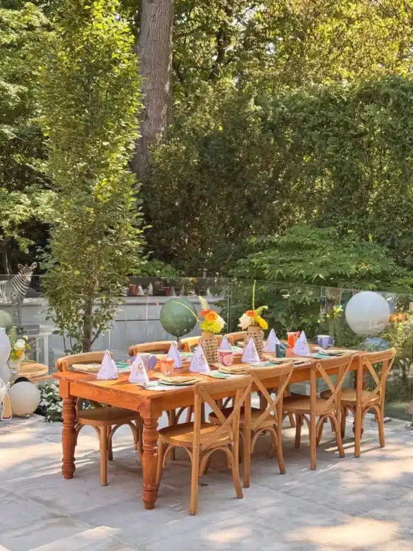 Children's safari theme 1st birthday table setting in Brampton with a large wooden harvest table and cross-back chairs on a patio, featuring matching party hats and floral centerpieces.