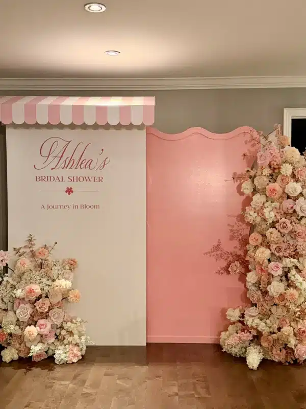 Custom pink wavy backdrop panel alongside a white backdrop with a pink striped awning, featuring a custom "Ashlea's Bridal Shower" decal and cascading pink and cream floral arrangements set up in a home in Mississauga.