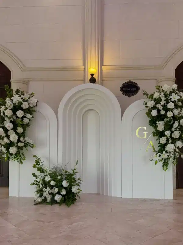 Three-piece white arched backdrop featuring a layered center ripple arch, decorated with white floral cascades and gold monogram lettering for a bridal shower at the Bellvue Event Centre.