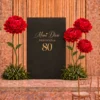 Black and gold birthday backdrop with custom gold lettering, oversized red flowers, tall green stems, and lush greenery arrangements for an elegant celebration.